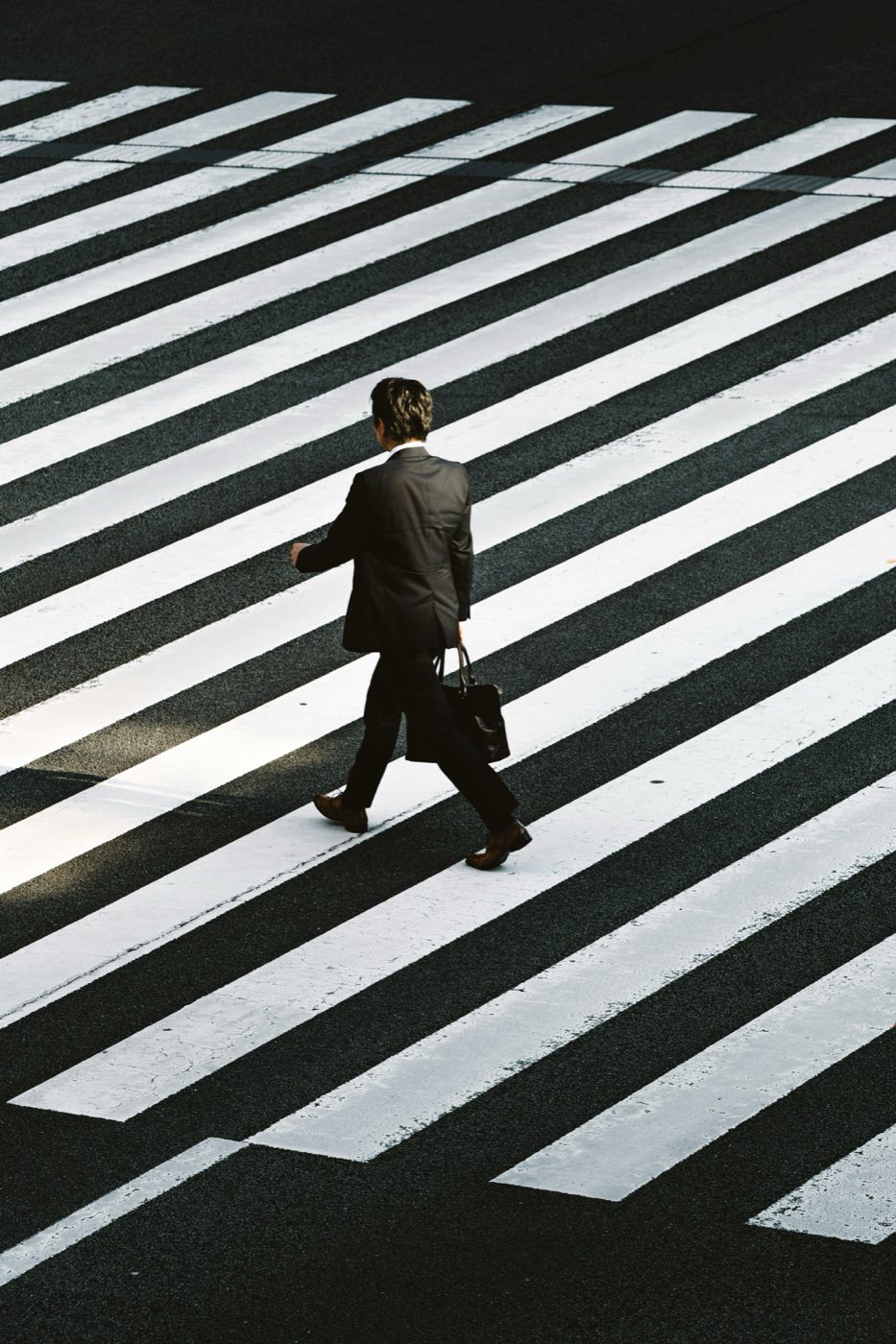 A single figure in a suit crossing a wide painted crosswalk, seen from above.